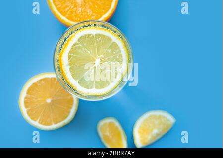 translucent glass of water with gases and bubbles, with a round slice of lemon on a glass and a whole lemon on blue background. Refreshment in the hea Stock Photo