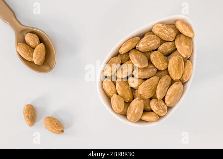 A white bowl filled with peeled almond seeds. A spoon with three peeled almond seeds and two more seeds, all on a white surface Stock Photo
