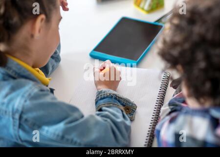 Boy and girl sitting at table with tablet, writing in their notebooks Stock Photo