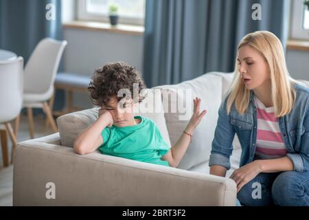 Sad curly boy sitting on sofa, blonde female talking to him, trying to cheer him up Stock Photo