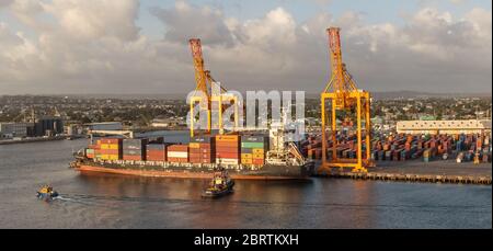 Bridgetown port with loading cranes and full cargo ship preparing to set sail. Tugboats ready to assist. Stock Photo