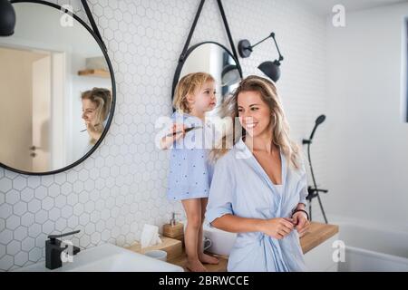 Young mother with small daughter indoors in bathroom, combing hair.