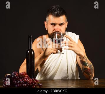 Sommelier smells drink. God Bacchus with serious face wearing white cloth sits by wine bottle and grapes. Man with beard holds glass of wine on brown background. Winemaking and degustation concept Stock Photo