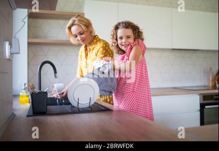 A cute small girl with mother indoors in kitchen at home, washing up dishes. Stock Photo