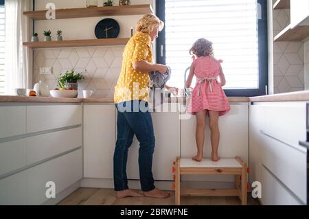 A rear view of small girl with mother indoors in kitchen, washing up dishes. Stock Photo