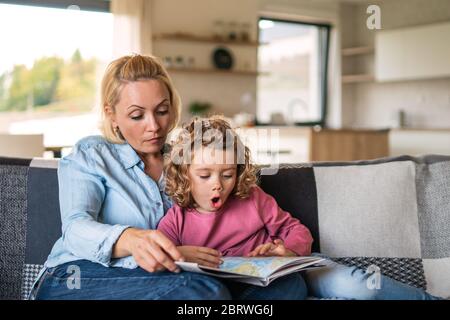 A cute small girl with mother on sofa indoors at home, reading book. Stock Photo