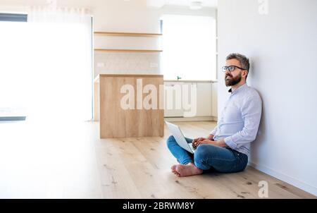 A mature man sitting on the floor in unfurnished new house, using laptop. Stock Photo