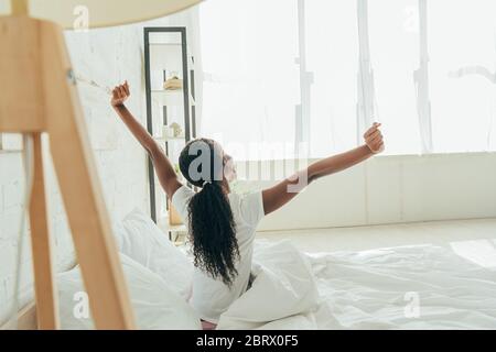back view of african american girl stretching with raised hands while sitting in bed Stock Photo