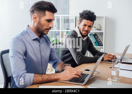 Selective focus of confused african american businessman looking at colleague using laptop in office Stock Photo