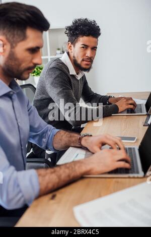 Selective focus of skeptical african american businessman looking at colleague using laptop near papers in office Stock Photo