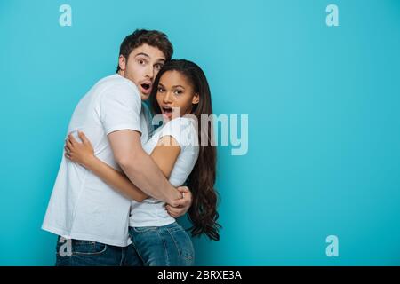 scared interracial couple embracing and looking at camera on blue background Stock Photo