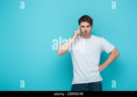 displeased man holding had on hip while talking on smartphone on blue background Stock Photo