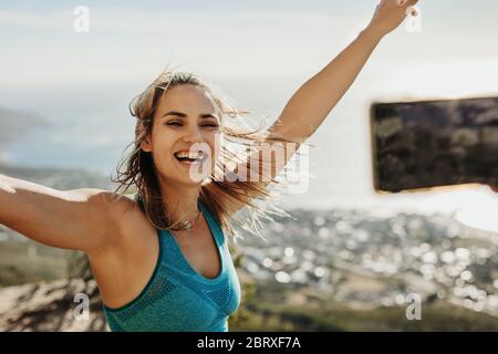 Excited woman being photographed by someone with a mobile phone on mountain top. Friend taking photos of a woman hiker in sportswear on mountain top w Stock Photo