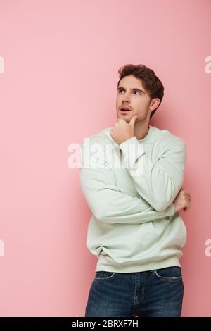 dreamy young man touching chin while looking away on pink background Stock Photo