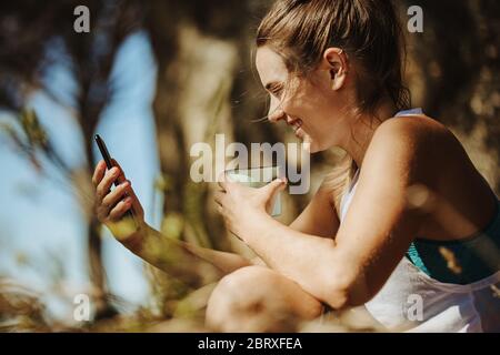 Smiling young woman looking at her phone and drinking coffee outdoors while on trekking. Sporty woman taking a break while hiking workout. Stock Photo