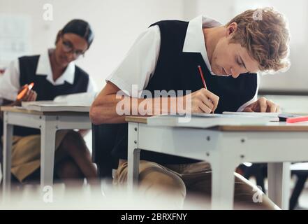 Teenage boy is taking a test while sitting at desk in classroom in high school. Students are concentrating while writing during exam. Stock Photo
