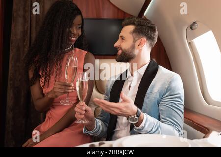 smiling interracial couple travelling in private jet and talking while holding glasses of champagne Stock Photo