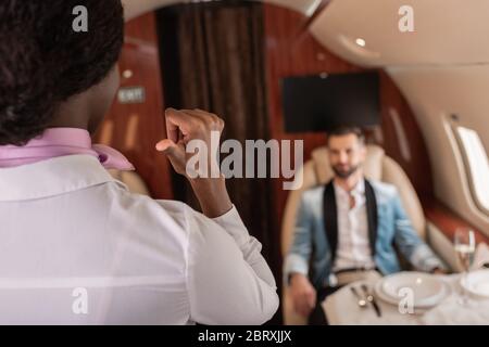 back view of african american stewardess pointing with thumb and elegant man sitting at served table in private plane Stock Photo