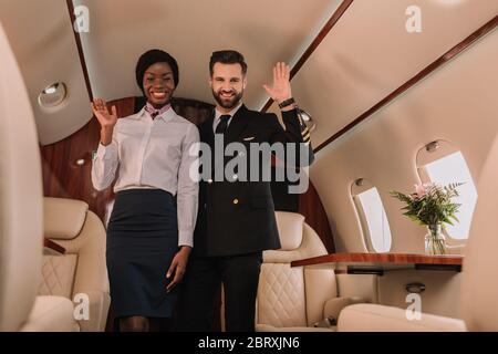 happy pilot and smiling afican american stewardess waving hands while looking at camera in private jet Stock Photo