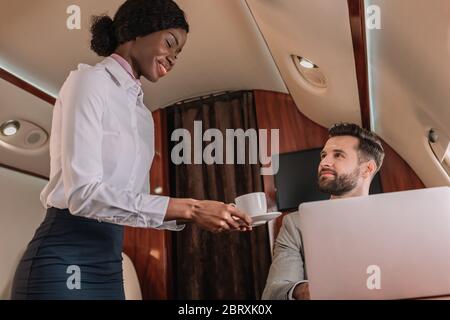 smiling african american stewardess giving cup of coffee to handsome businessman working on laptop in private jet Stock Photo