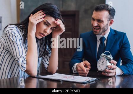 Angry collector holding alarm clock and pointing at documents near stressed woman at table in room Stock Photo