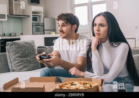 KYIV, UKRAINE - APRIL 16, 2020: sad girl sitting near boyfriend playing video game with joystick during self isolation at home with pizza Stock Photo