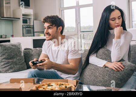 KYIV, UKRAINE - APRIL 16, 2020: offended woman sitting near concentrated man playing video game with joystick during self isolation at home with pizza Stock Photo