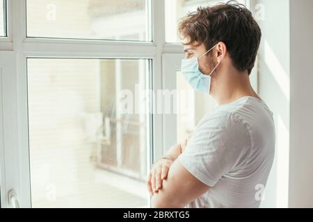 sad ill man in medical mask with crossed arms looking through window during self isolation Stock Photo