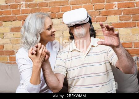 happy and mature woman looking at husband in virtual reality headset gesturing at home Stock Photo