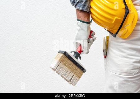 Caucasian house painter worker in white work overalls and with protective gloves, he holds the brush in his hand. Helmet and protective goggles. Work Stock Photo