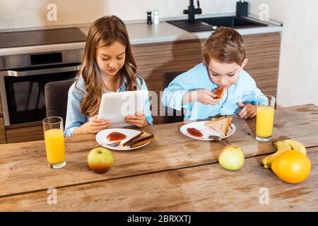 boy eating toast bread with jam near sister using digital tablet Stock Photo