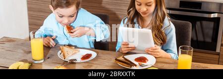 panoramic crop of boy eating toast bread with jam near sister using digital tablet Stock Photo