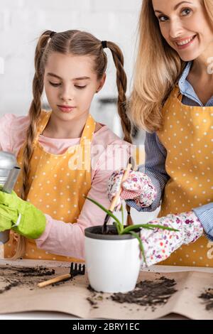 Woman with shovel and flowerpot with aloe smiling and cute daughter holding watering pot near table in kitchen Stock Photo
