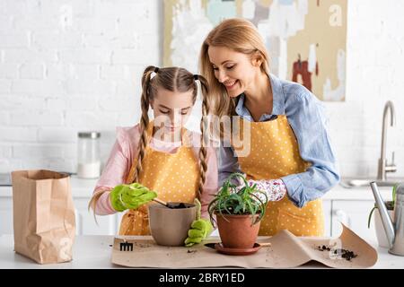 Woman smiling near cute daughter holding shovel with ground in flowerpot near table in kitchen Stock Photo
