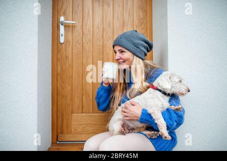 Mature woman relaxing outdoors by front door at home with coffee and pet dog. Stock Photo