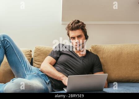 serious man in headset lying on sofa near laptop and looking at camera Stock Photo