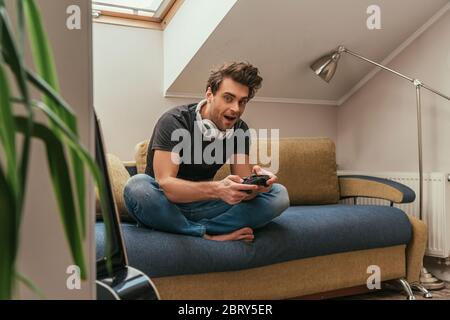 KYIV, UKRAINE - APRIL 13, 2019: selective focus of excited man in wireless headphones on neck playing video game and looking at camera Stock Photo