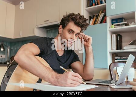 handsome young man with guitar writing notes and looking at camera while sitting near laptop Stock Photo