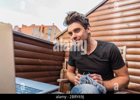 attentive young man looking at laptop while knitting on balcony Stock Photo