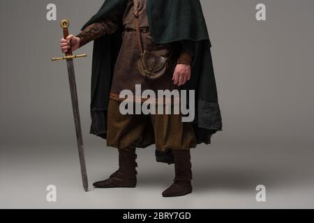 cropped view of medieval Scottish knight in mantel with sword on grey background Stock Photo