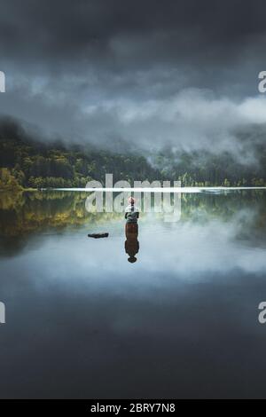 Amazing rear view reflection of a man standing on rock in the middle of a lake. Folk scenery concept with cloudy sky and and stunning fog conditions r Stock Photo
