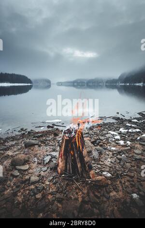 Small campfire with gentle flames beside a lake during a winter on a moody morning, Firiza, Maramures, Romania Stock Photo