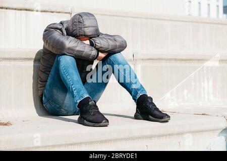 a young caucasian man, wearing jeans and a gray hooded jacket, curled up sitting on an outdoor concrete stairway Stock Photo