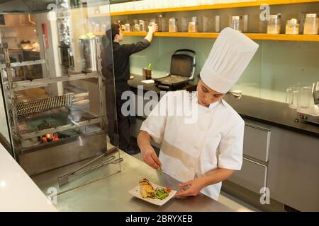 Female chef wearing chef's hat standing at worktop in commercial  kitchen, garnishing plate of food. Stock Photo