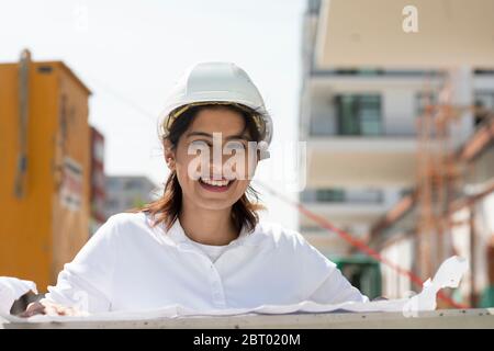 Female architect wearing white hard hat working on construction site, smiling at camera. Stock Photo