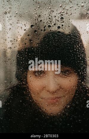Head and shoulders portrait of woman wearing knit hat, looking at camera through window covered in raindrops. Stock Photo
