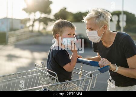 Mother joyfully playing with kid sitting in shopping cart. Both wearing protective face mask before entering a supermarket.