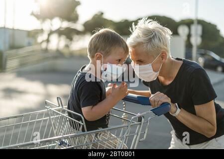 Mother joyfully playing with kid sitting in shopping cart. Both wearing protective face mask before entering a supermarket.