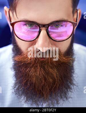 Bearded man in glasses with screen reflection working on computer in IT office, sitting at desk writing code, working on a project in software Stock Photo