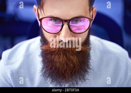 Bearded man in glasses with screen reflection working on computer in IT office, sitting at desk writing code, working on a project in software Stock Photo